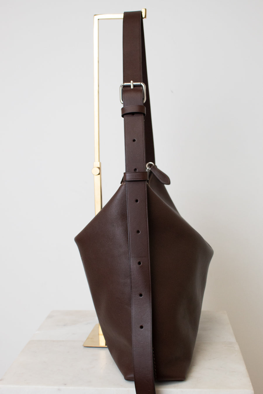 A dark brown leather bucket bag with a wide adjustable shoulder strap, displayed on a minimalist gold metal stand against a light gray background.