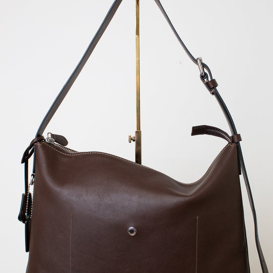  A dark brown leather shoulder bag with a soft, slouchy silhouette with a adjustable strap with silver hardware , the bag is photographed upright on a white background hanging from a gold hardware stand.