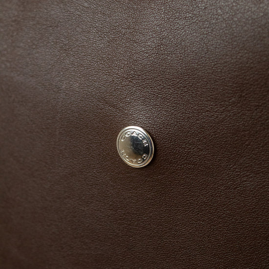 A close-up of  a dark brown textured leather Coach bag , featuring a small round silver toned metal emblem centered on the  surface with COACH engraved in it.