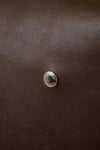 A close-up of  a dark brown textured leather Coach bag , featuring a small round silver toned metal emblem centered on the  surface with COACH engraved in it.