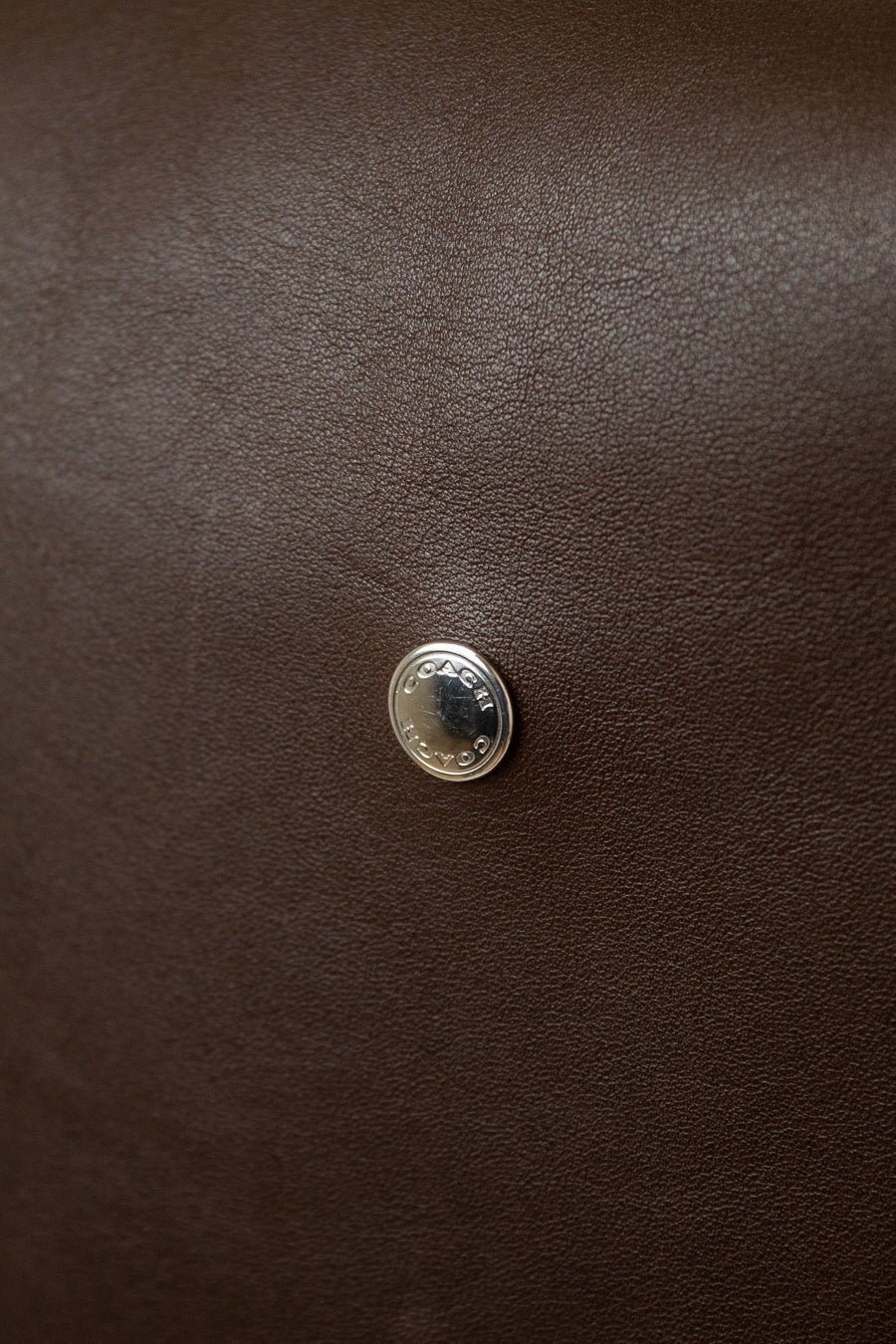 A close-up of  a dark brown textured leather Coach bag , featuring a small round silver toned metal emblem centered on the  surface with COACH engraved in it.