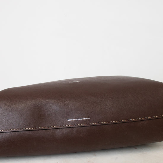 A close - up of a dark brown leather bag showing a smooth,slightly glossy surface. A horizontal stitch seam runs across the lower portion of the leather with neat evenly spaced out brown stitching. 