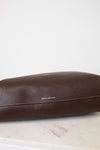 A close - up of a dark brown leather bag showing a smooth,slightly glossy surface. A horizontal stitch seam runs across the lower portion of the leather with neat evenly spaced out brown stitching. 