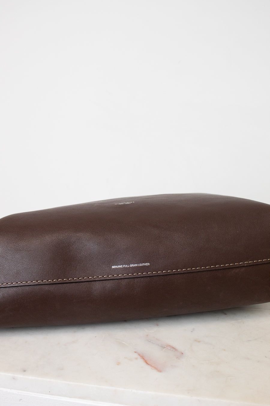 A close - up of a dark brown leather bag showing a smooth,slightly glossy surface. A horizontal stitch seam runs across the lower portion of the leather with neat evenly spaced out brown stitching. 