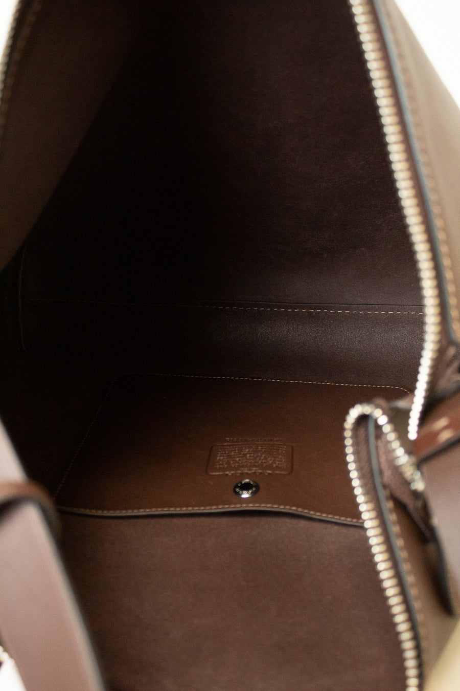 Close-up of the interior of a brown leather handbag, showing the dark brown lining, silver-tone zipper, and a small embossed brand patch on the bottom.