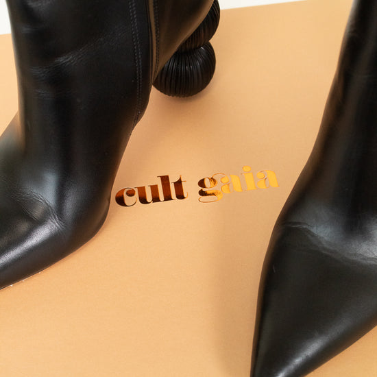 Black leather ankle boots with pointed toes and stacked block heels, displayed on a beige box with the brand name 'cult gaia' printed on it.