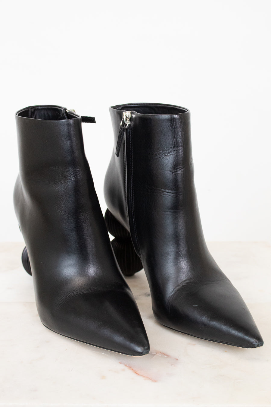 A pair of black leather pointed-toe ankle boots with side zippers, displayed on a light marble surface against a white background.