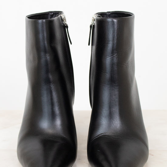 A pair of black leather pointed-toe ankle boots with side zippers, displayed on a light marble surface against a white background.