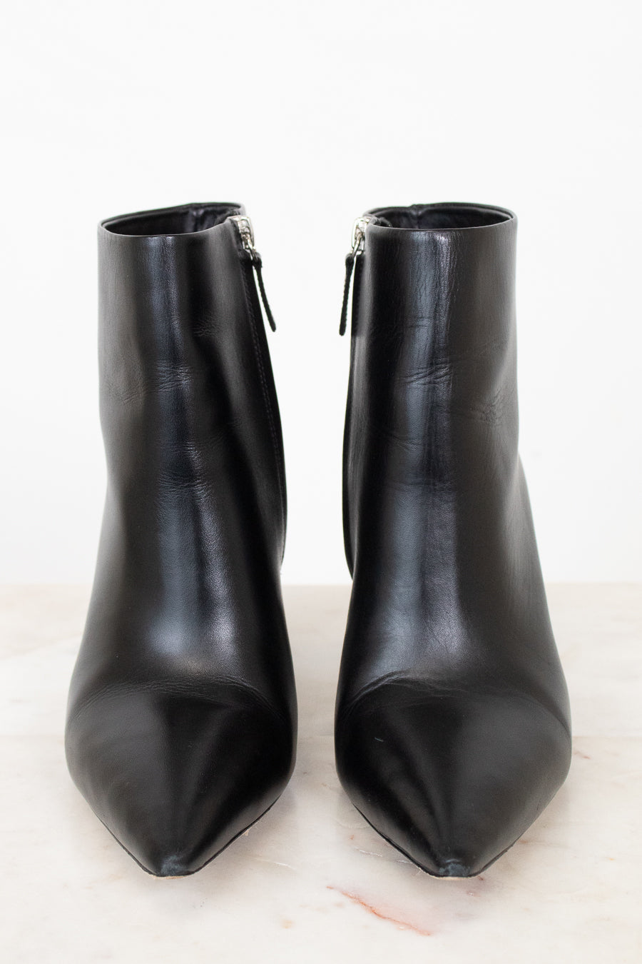 A pair of black leather pointed-toe ankle boots with side zippers, displayed on a light marble surface against a white background.