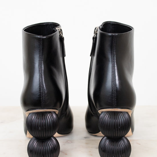 A pair of black leather ankle boots with distinctive stacked spherical block heels, shown from the back on a light marble surface against a white background.
