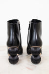 A pair of black leather ankle boots with distinctive stacked spherical block heels, shown from the back on a light marble surface against a white background.