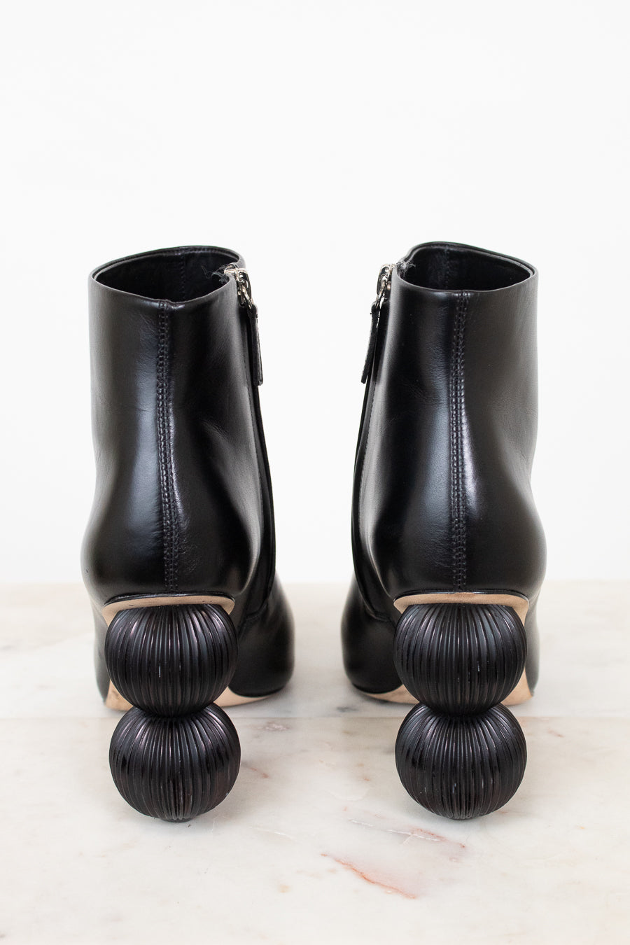 A pair of black leather ankle boots with distinctive stacked spherical block heels, shown from the back on a light marble surface against a white background.