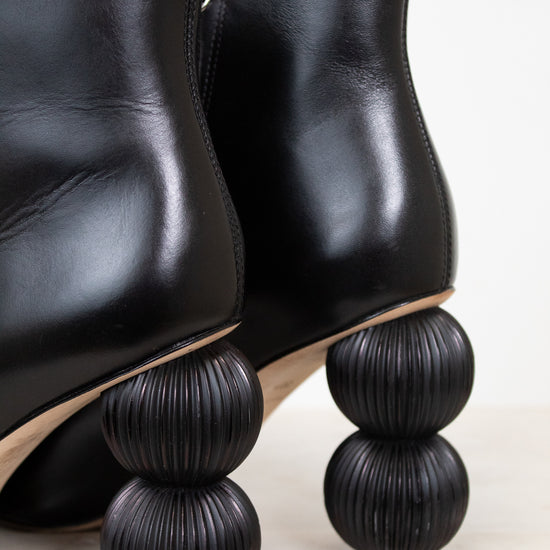 Black leather ankle boots with distinctive stacked spherical black heels, shown on a light marble surface against a white background.