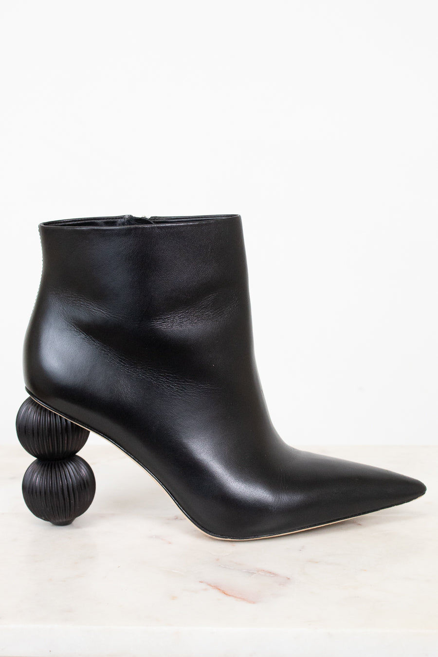 A black leather ankle boot with a pointed toe and a unique stacked spherical heel, displayed on a light marble surface against a white background.