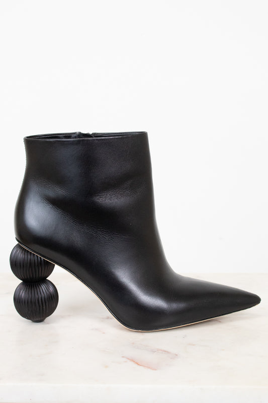 A black leather ankle boot with a pointed toe and a unique stacked spherical heel, displayed on a light marble surface against a white background.