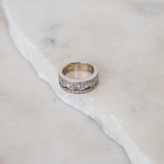 A silver band ring with a row of clear diamonds, resting on a white marble slab against a light marble background.