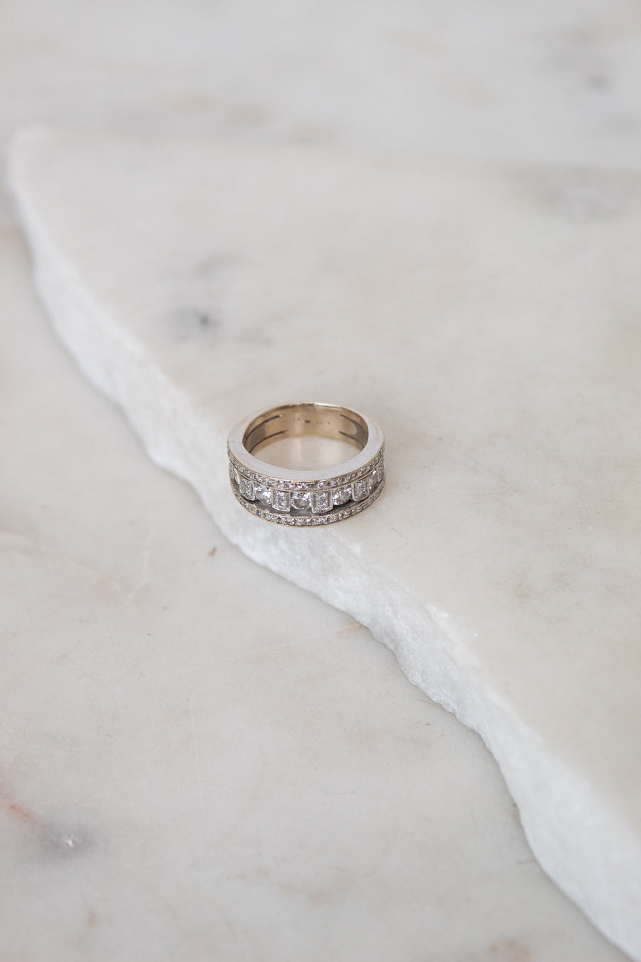 A silver band ring with a row of clear diamonds, resting on a white marble slab against a light marble background.