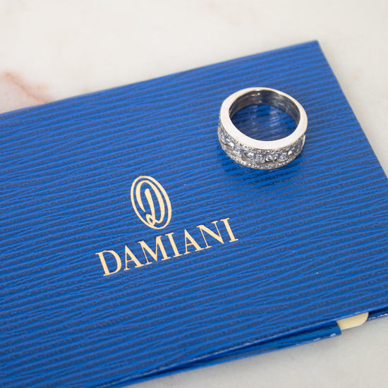 A silver ring with diamonds rests on a textured blue Damiani box, placed on a light marble surface.