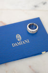 A silver ring with diamonds rests on a textured blue Damiani box, placed on a light marble surface.