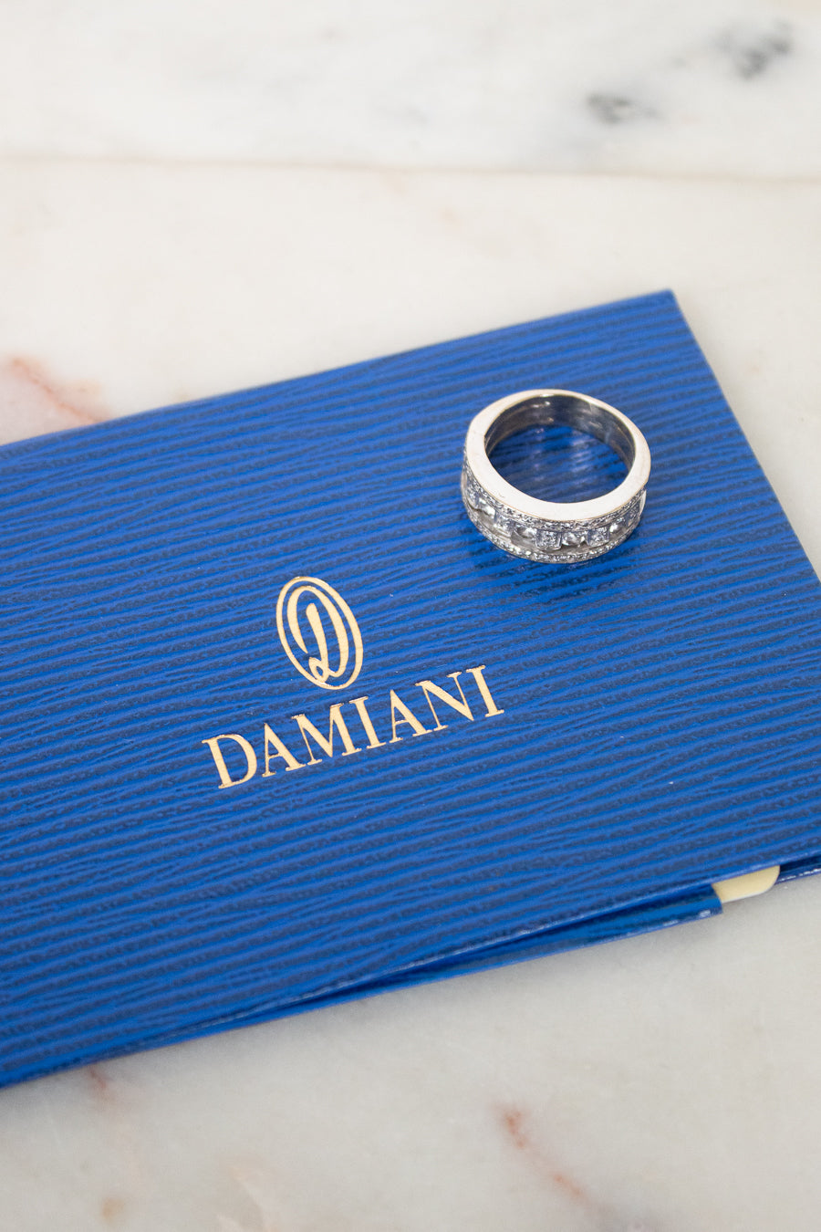 A silver ring with diamonds rests on a textured blue Damiani box, placed on a light marble surface.