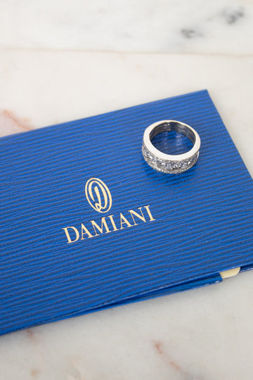 A silver ring with diamonds rests on a textured blue Damiani box, placed on a light marble surface.