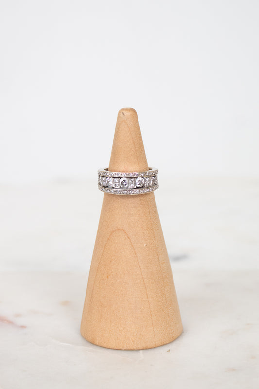 A silver band ring set with diamonds, displayed on a light wood cone stand against a white background.