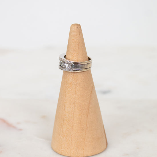 A silver band ring with diamonds set on a light natural wood cone ring holder, placed on a white marble surface against a white background.