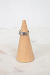 A silver band ring with diamonds set on a light natural wood cone ring holder, placed on a white marble surface against a white background.