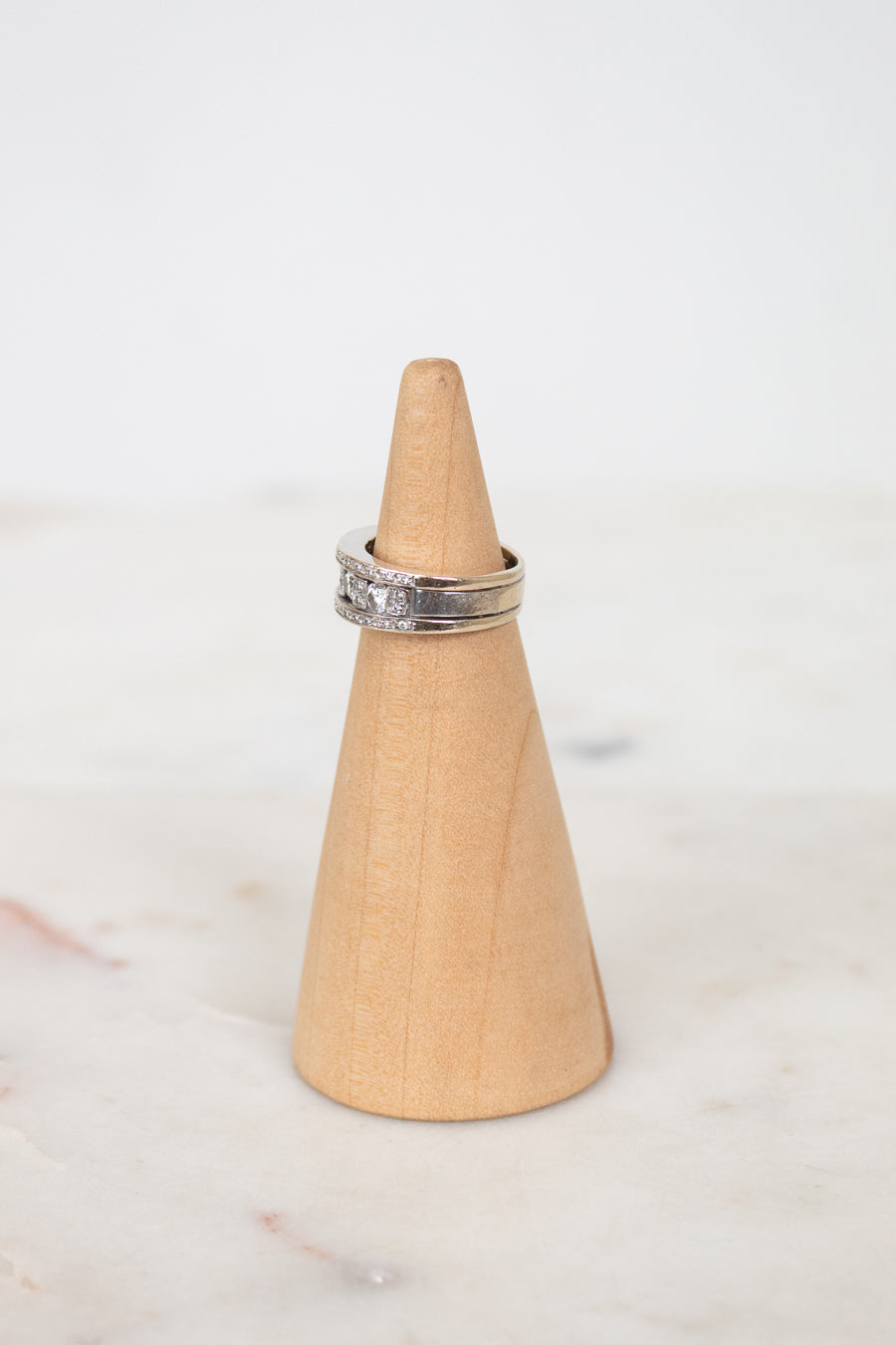 A silver band ring with diamonds set on a light natural wood cone ring holder, placed on a white marble surface against a white background.