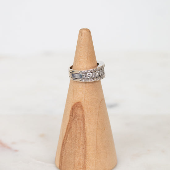 A silver band ring with diamonds, displayed on a light wood cone stand against a soft white background.