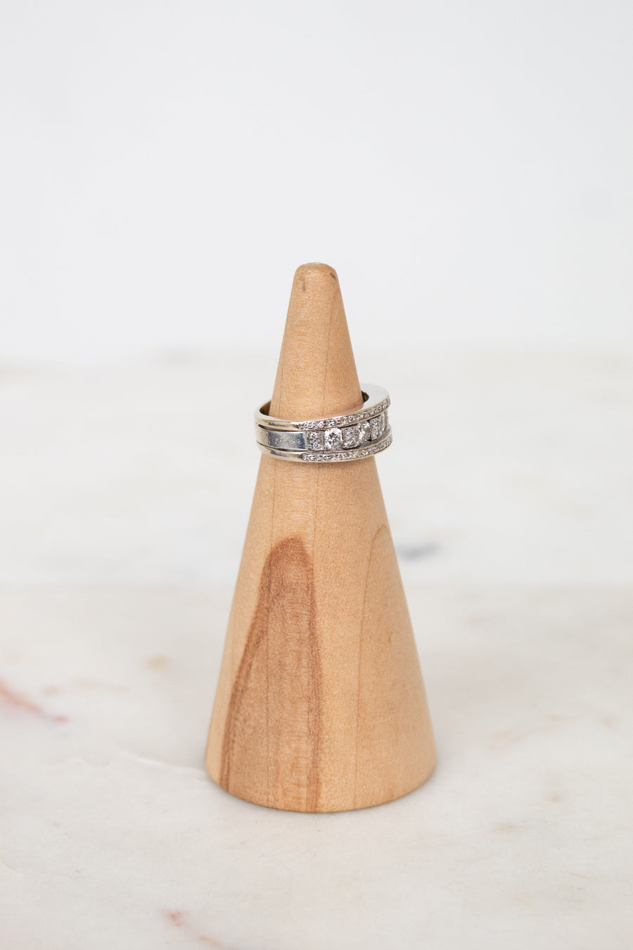 A silver band ring with diamonds, displayed on a light wood cone stand against a soft white background.