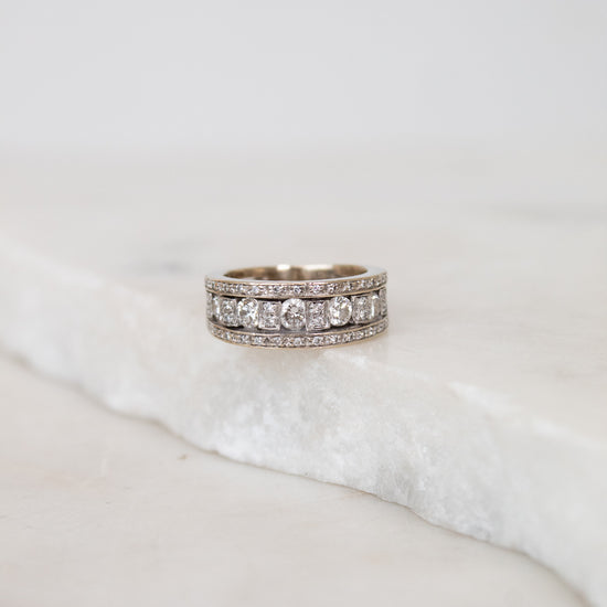 A silver band ring with multiple rows of diamonds, resting on a white marble slab against a soft light gray background.