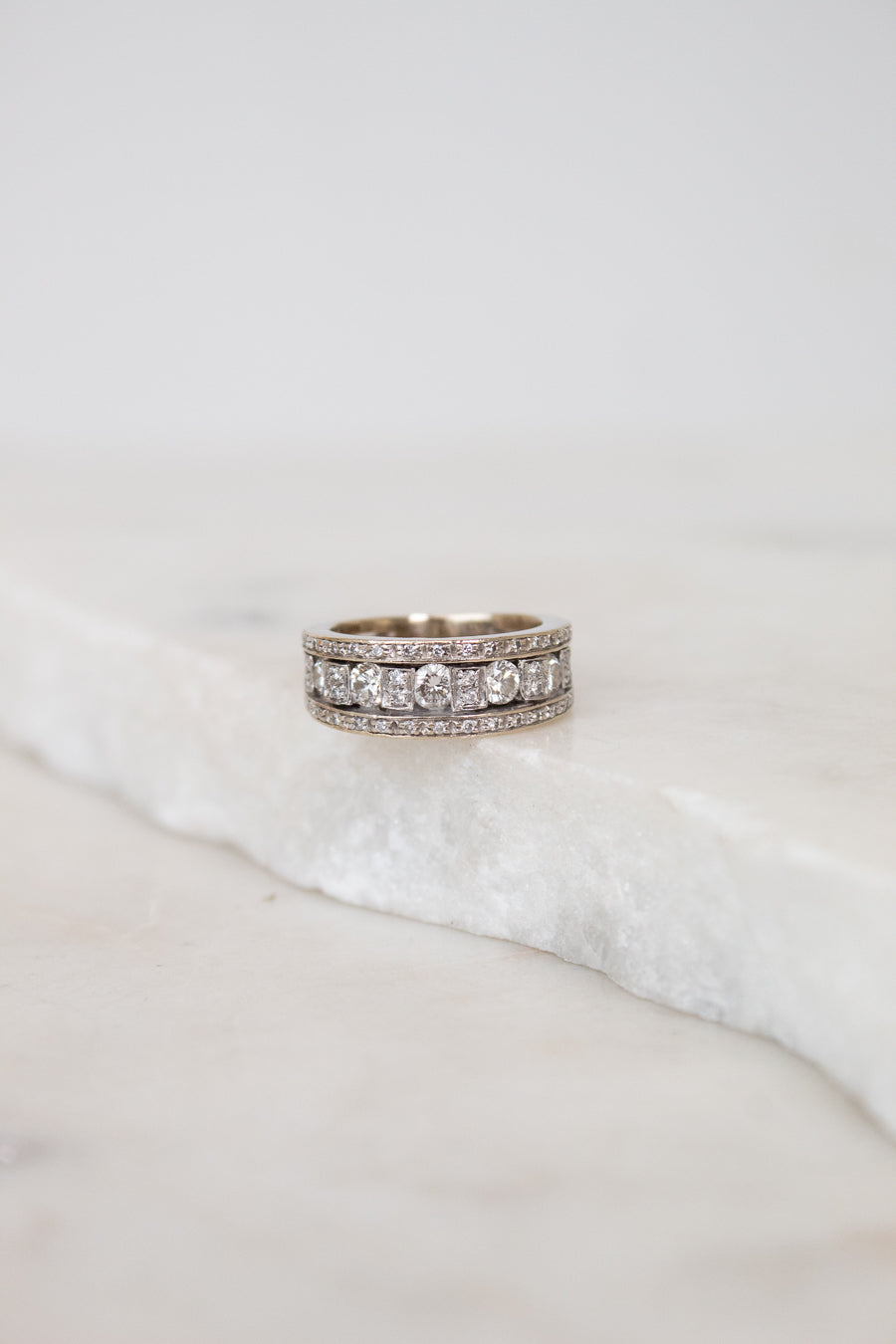 A silver band ring with multiple rows of diamonds, resting on a white marble slab against a soft light gray background.