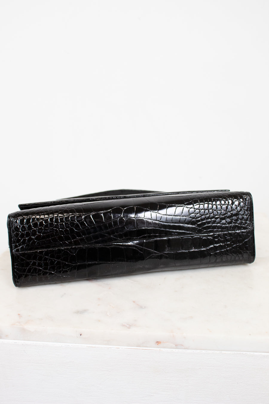 A black crocodile-embossed leather handbag, shown closed on a white marble surface against a white background.