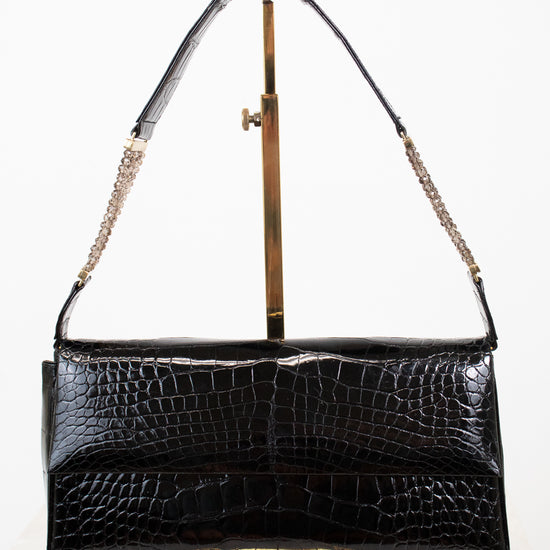 A black crocodile-embossed leather shoulder bag with gold-tone hardware, displayed on a gold metal stand against a white background.