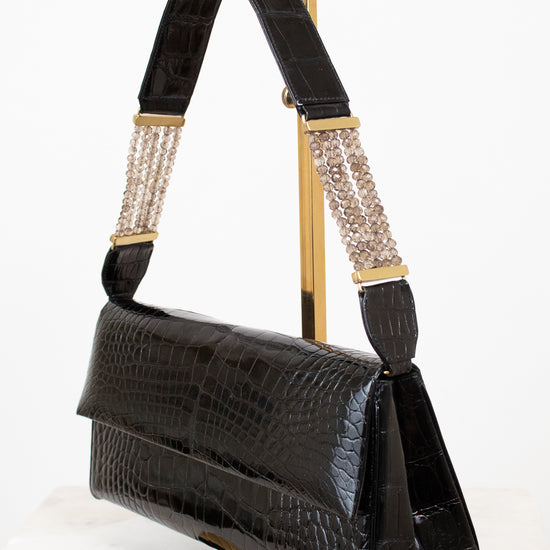 Black crocodile-embossed leather shoulder bag with gold-tone hardware and a beaded chain detail on the strap, displayed on a gold stand against a white background.