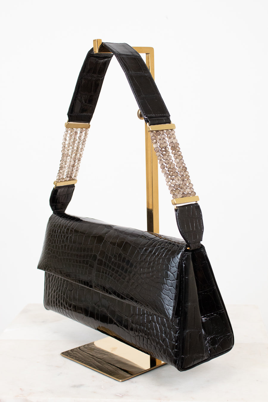 Black crocodile-embossed leather shoulder bag with gold-tone hardware and a beaded chain detail on the strap, displayed on a gold stand against a white background.
