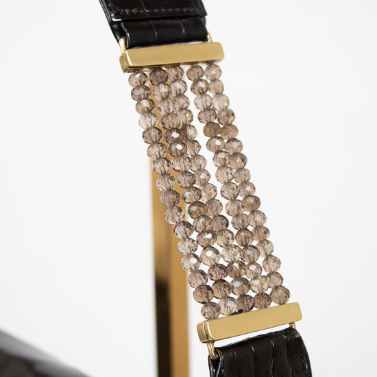 Close-up of a black crocodile-embossed shoulder strap with gold-tone hardware and a central cluster of faceted champagne-beige beads, shown against a white background.
