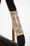 Close-up of a black crocodile-embossed shoulder strap with gold-tone hardware and a central cluster of faceted champagne-beige beads, shown against a white background.
