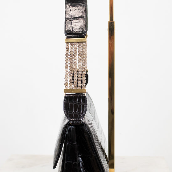 A black crocodile-embossed leather handbag with a wide, decorative strap featuring gold-tone hardware and a row of clear rhinestones, displayed on a gold metal stand against a white background.
