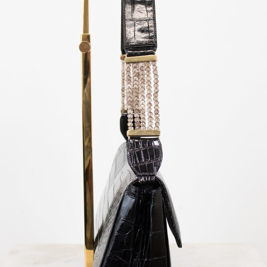 A black crocodile-embossed leather handbag with a gold-tone stand, featuring a wide strap with a woven beige-and-black center and gold-tone hardware, set against a plain white background.