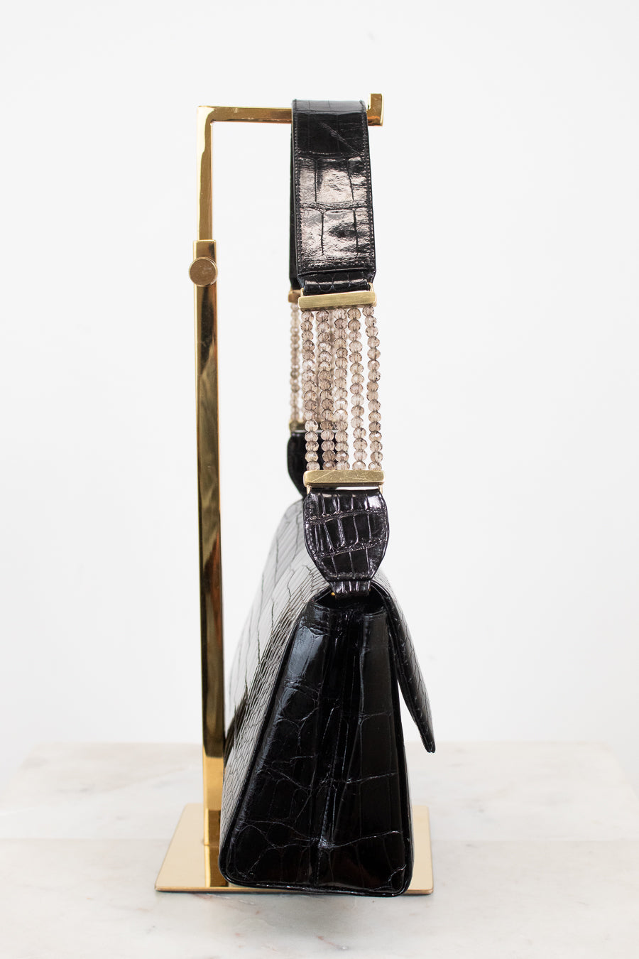 A black crocodile-embossed leather handbag with a gold-tone stand, featuring a wide strap with a woven beige-and-black center and gold-tone hardware, set against a plain white background.