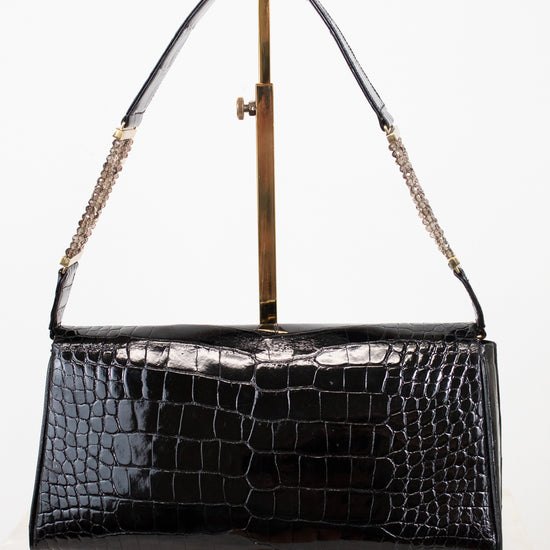 A black crocodile-embossed leather handbag with a gold-tone chain strap, displayed on a gold metal stand against a white background.