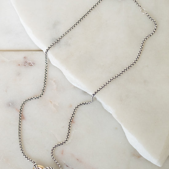 A delicate silver box chain necklace with a two-tone pendant featuring a gold and silver interlocked ring design, displayed on a white marble slab.