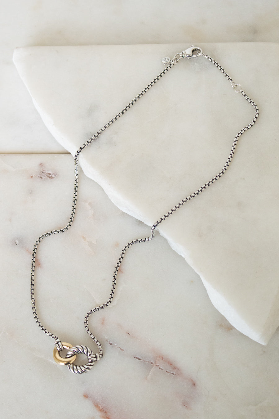 A delicate silver box chain necklace with a two-tone pendant featuring a gold and silver interlocked ring design, displayed on a white marble slab.