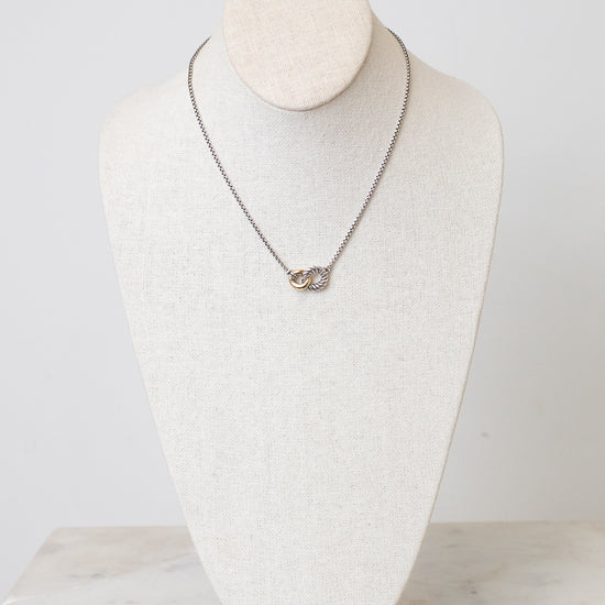 A silver chain necklace with a small interlocked rings pendant displayed on a cream-colored fabric jewelry bust against a light gray background.