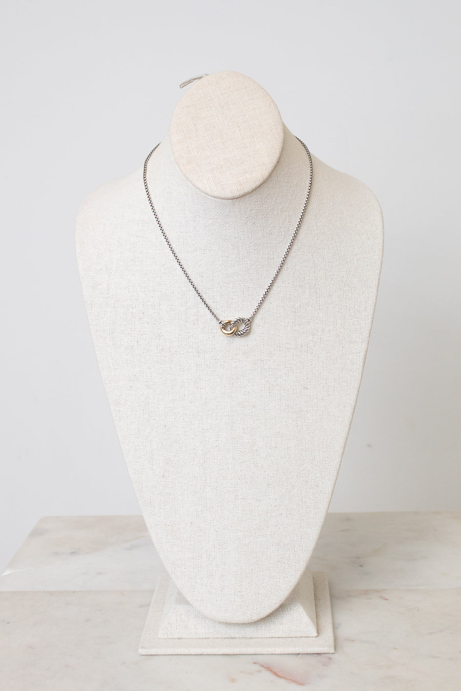 A silver chain necklace with a small interlocked rings pendant displayed on a cream-colored fabric jewelry bust against a light gray background.