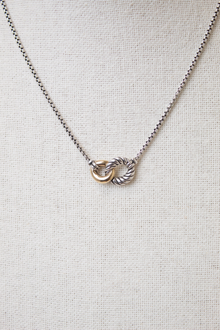 Interlocking gold and ribbed silver rings attached to dainty box chain necklace displayed against a plain textured background
