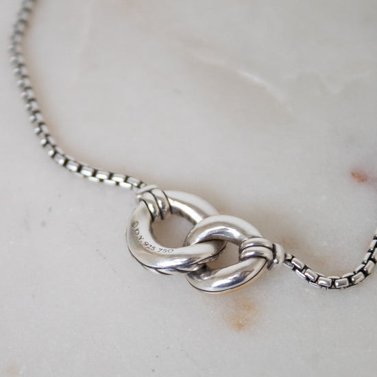 A close-up of a silver box chain necklace showing the back of an interlocked ring pendant, resting on a light beige marble surface.