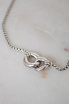 A close-up of a silver box chain necklace showing the back of an interlocked ring pendant, resting on a light beige marble surface.